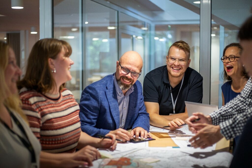 Ryhmä ihmisiä istuu lasiseinäisessä neuvotteluhuoneessa hymyillen ja keskustellen karttojen ja asiakirjojen äärellä.